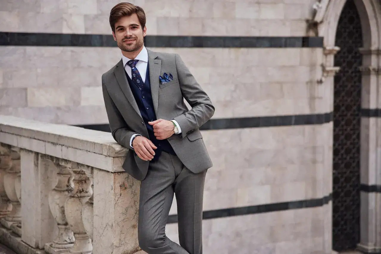 Elegancki mężczyzna w szarym garniturze, granatowej kamizelce i wzorzystym krawacie. Idealne buty do szarego garnituru? Zobacz inspirację.